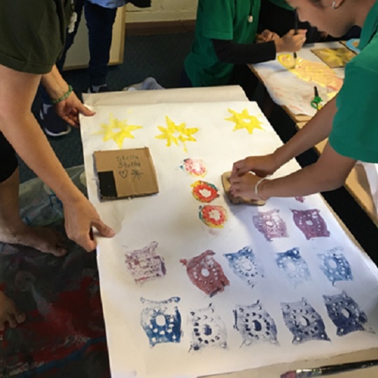 Photo of students and Martha block printing for the Mindfulness Art Project, image shows hands on paper block printing with shapes on a large piece of white paper